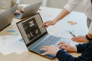 社員数名がテーブルに向かい合って座り画面手前の社員のPCのモニターに別の社員が指示を出している様子
